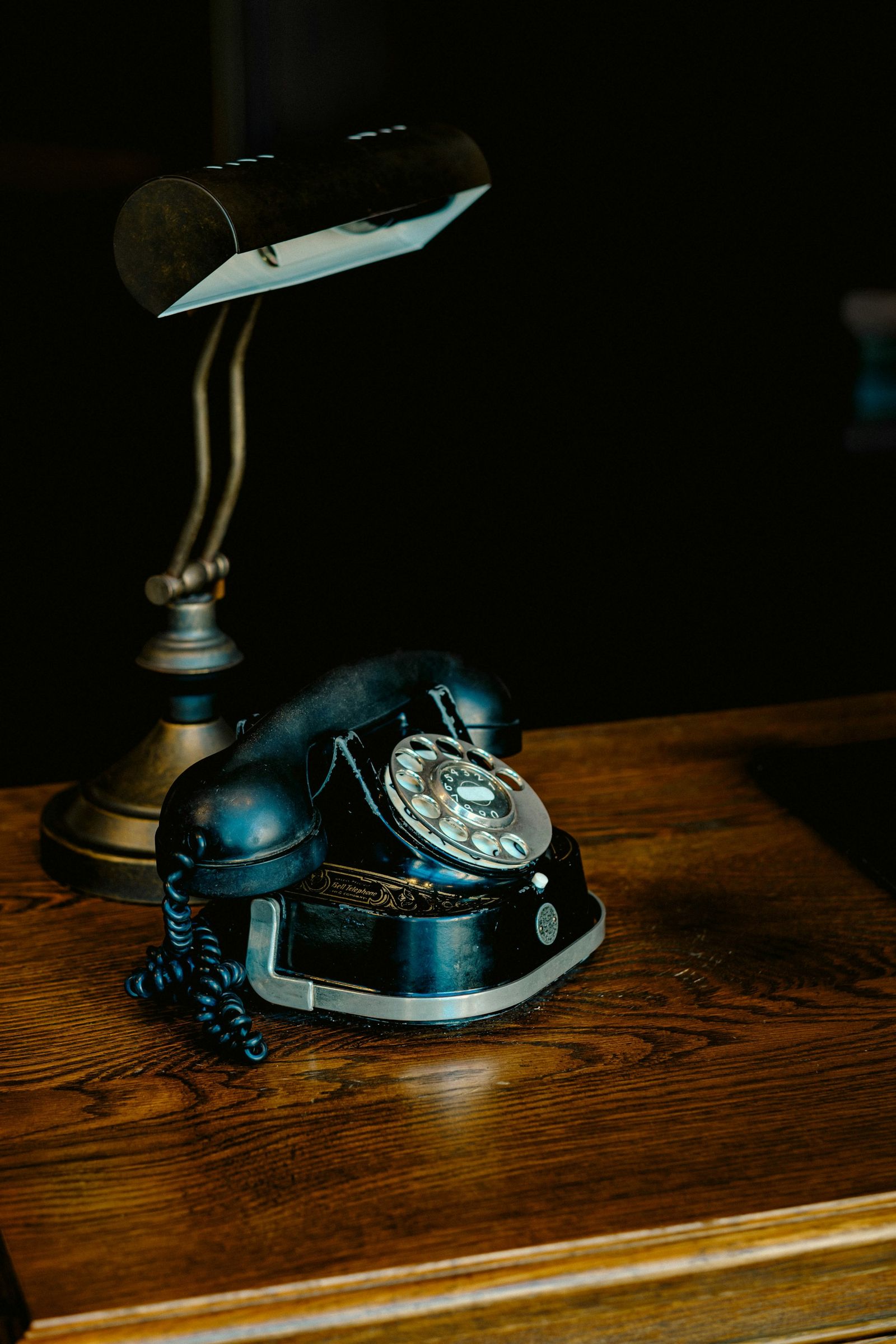 A vintage rotary telephone beside a brass desk lamp on a wooden desk in the evening